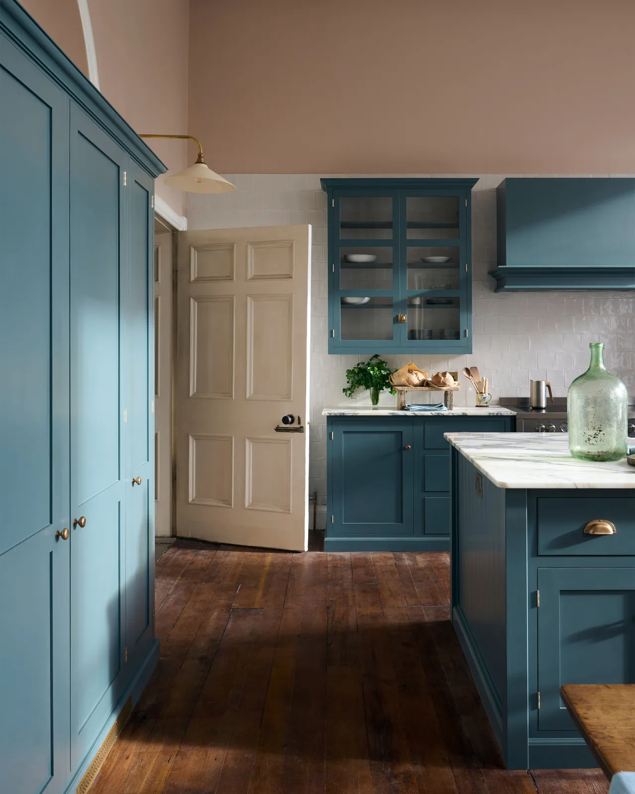 An impressive room with high ceilings, plaster pink walls, rich worn floorboards and a green-blue painted Shaker-style kitchen.