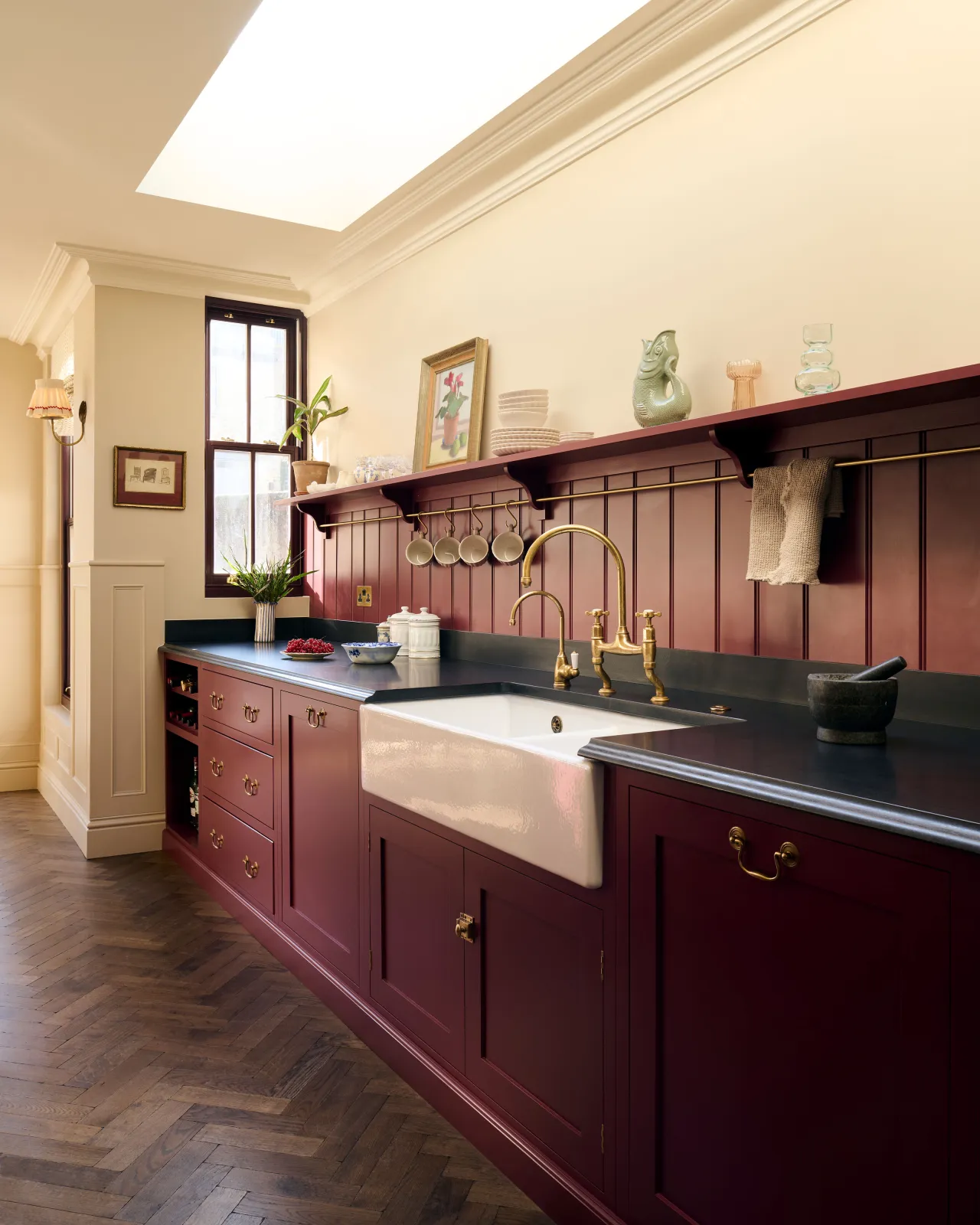 An oblique view of a red painted Shaker-style kitchen with parquet wood flooring, cream walls, a black granite worktop and red tongue and groove panelling above.