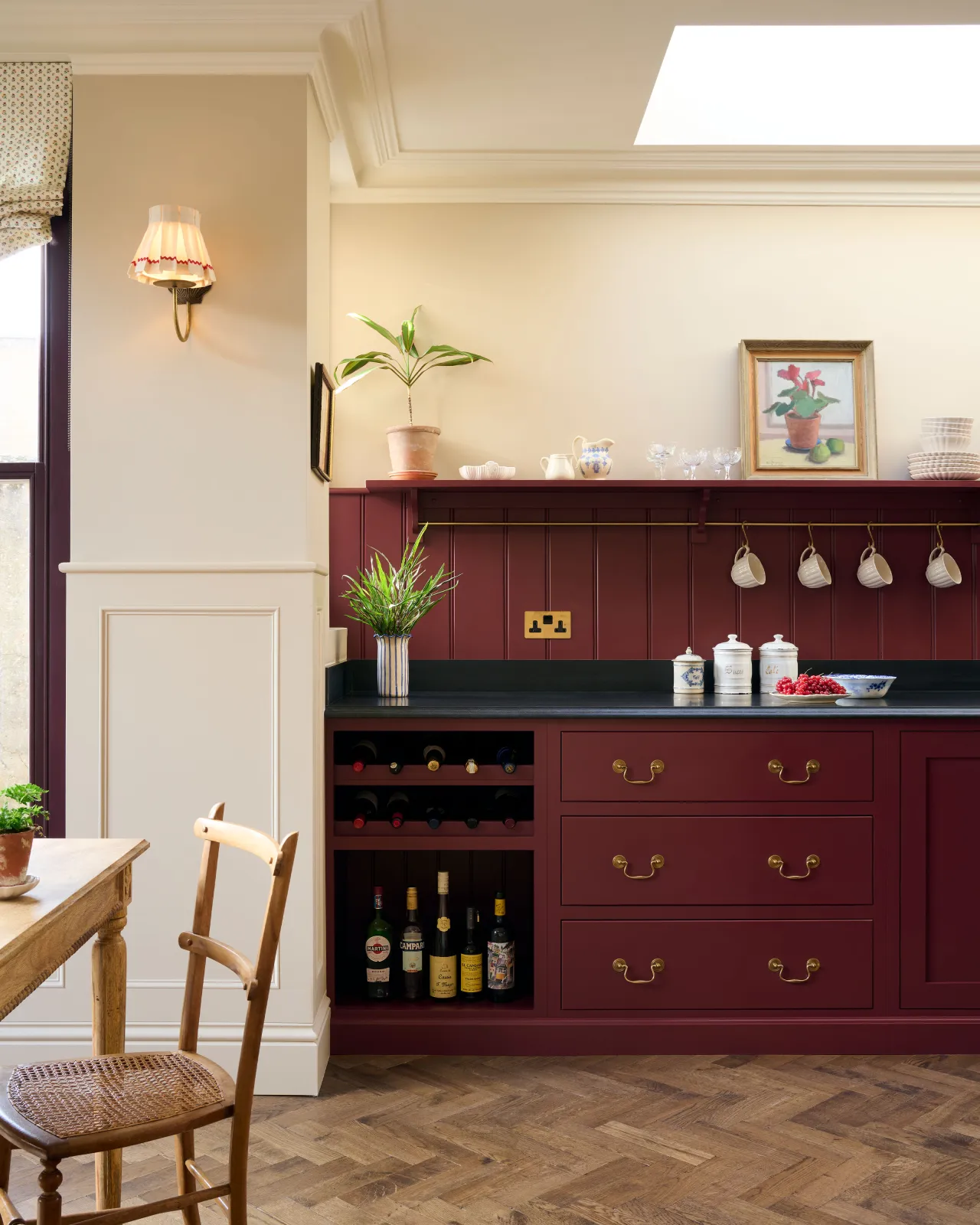 A straight-on view of a red painted Shaker-style kitchen with parquet wood flooring, cream walls, a black granite worktop and red tongue and groove panelling above.