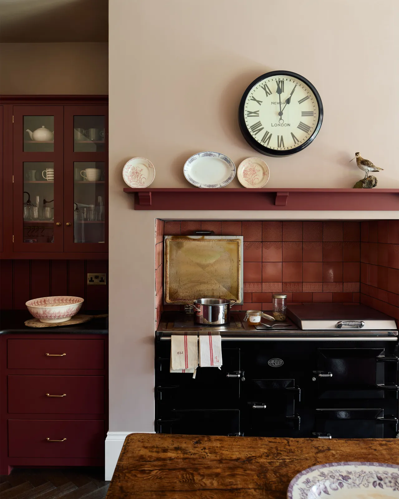 A straight-on view of a red painted Shaker-style kitchen with a large black Aga cooker in a chimney breast alcove with glossy red patterned wall tiles above.