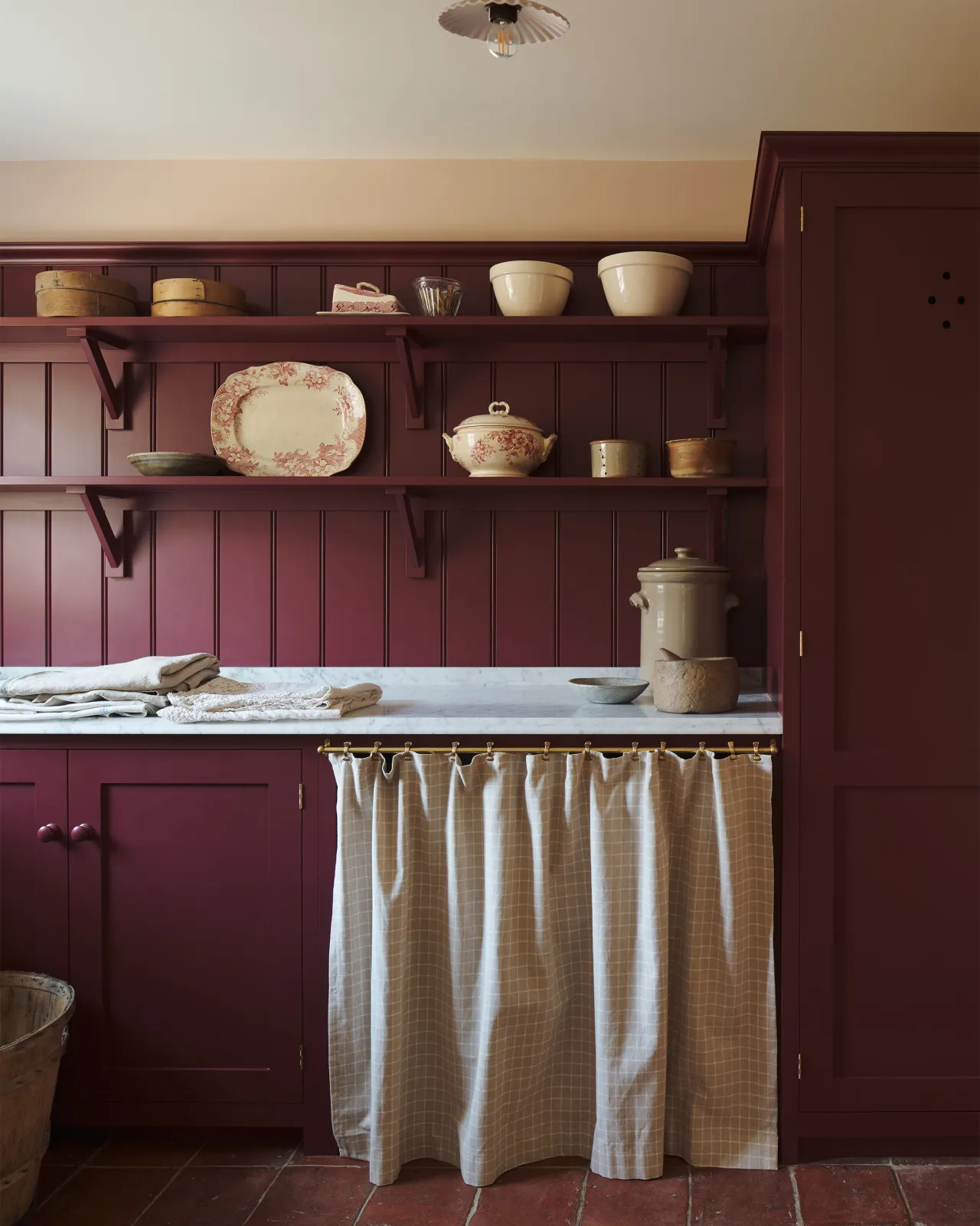 A straight-on view of a red painted Shaker-style utility kitchen with a tall pantry cupboard, open storage behind a red striped curtain and tongue and groove wall panelling and shallow shelves decorated with vintage crockery.