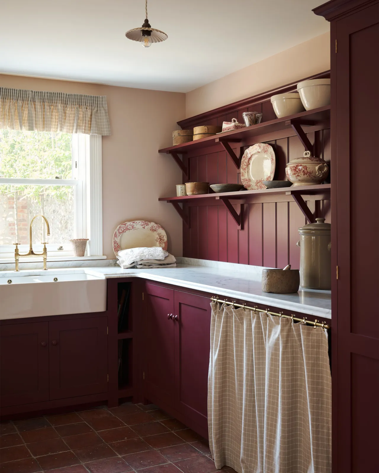 An oblique view of a red painted Shaker-style utility kitchen with a tall pantry cupboard, open storage behind a red striped curtain and tongue and groove wall panelling and shallow shelves decorated with vintage crockery.