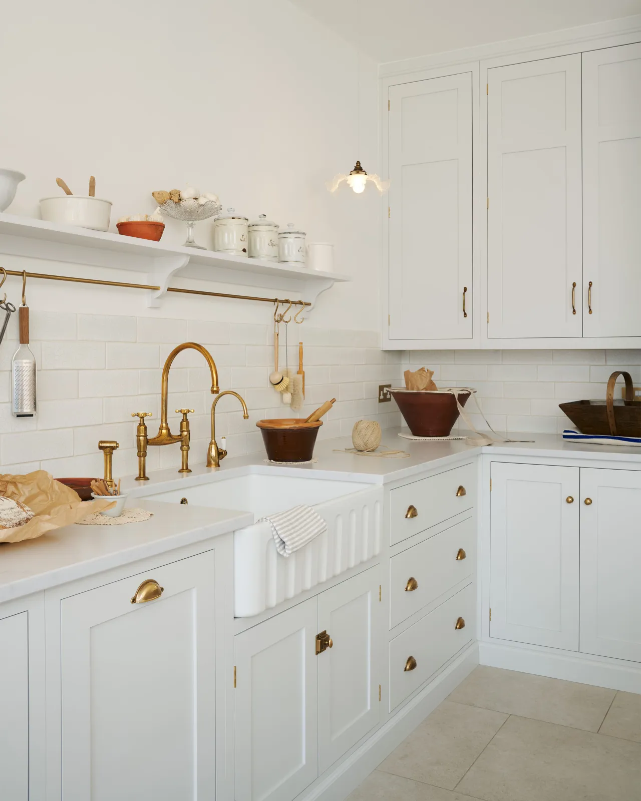 An oblique view of a simple Shaker-style kitchen with off-white painted cupboards, a fluted Belfast sink, aged brass taps and white metro tiles.