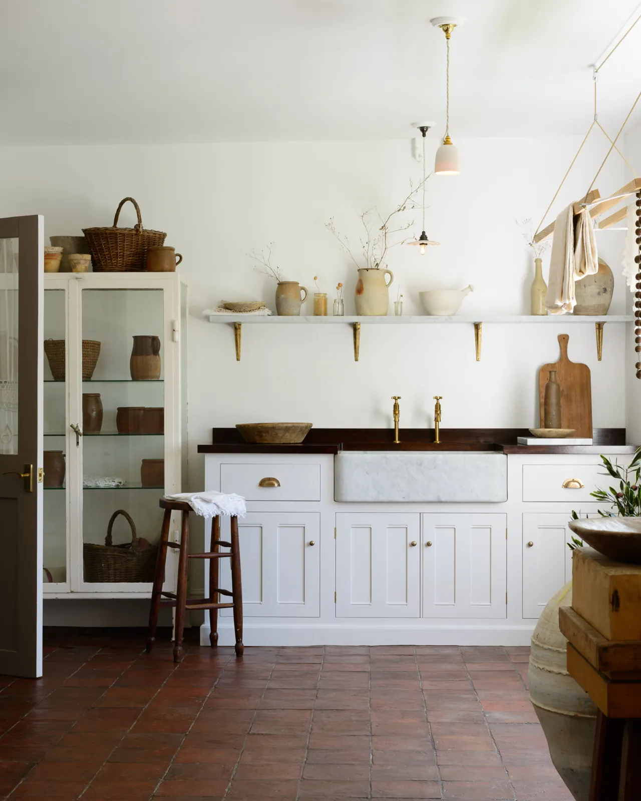 A Mediterannean-inspired scullery room with white walls, a worn terracotta floor and an off-white painted sink cabinet with a dark hardwood worktop, aged brass taps and a Carrara marble sink.