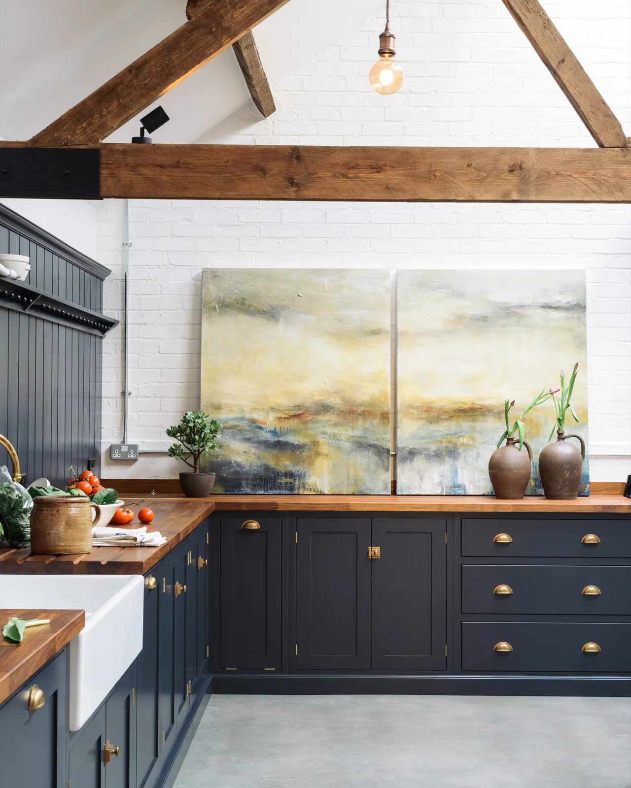 A large barn conversion with a simple Shaker-style kitchen, dark blue painted cupboards, brass hardware, rich hardwood worktops, white brick walls, a cement floor and old worn ceiling beams.