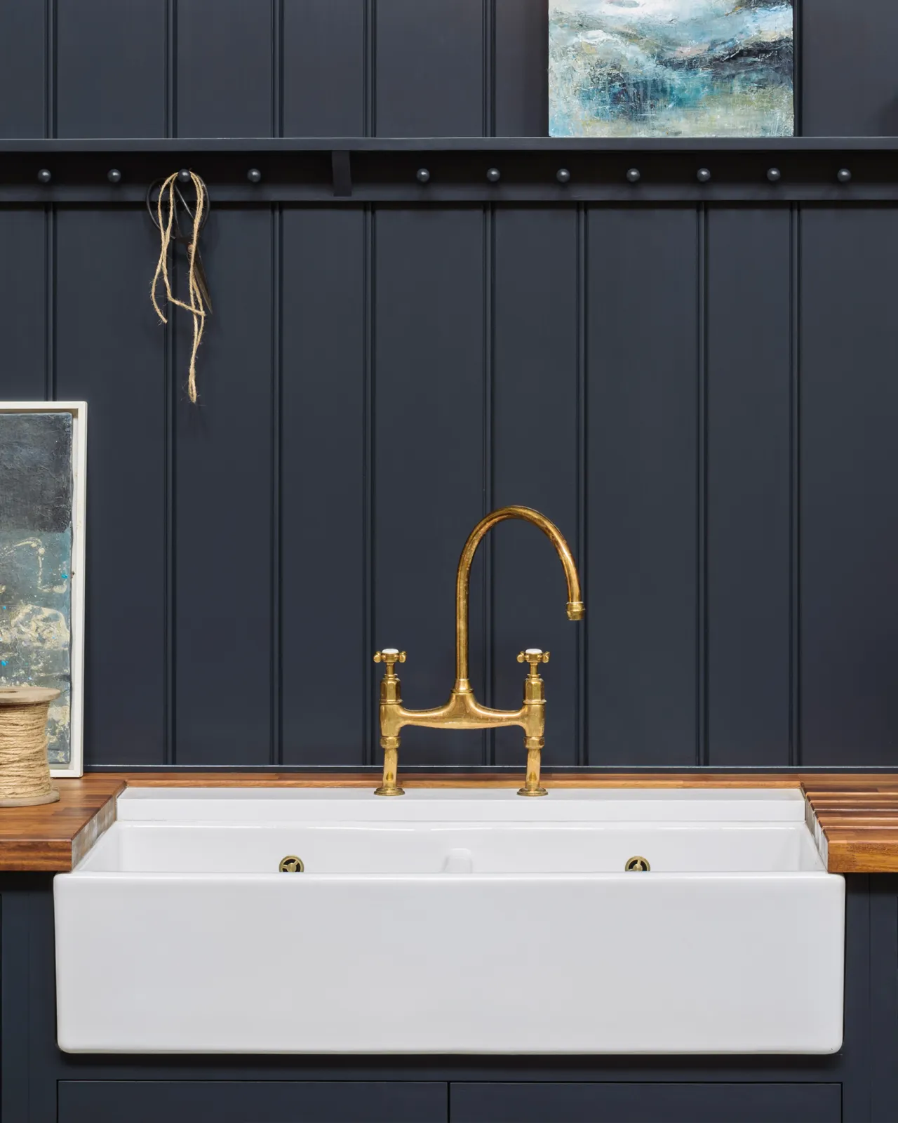 A close-up view of a wide ceramic Belfast sink with an aged brass tap, rich hardwood worktop and dark blue painted tongue and groove-style panelling behind.