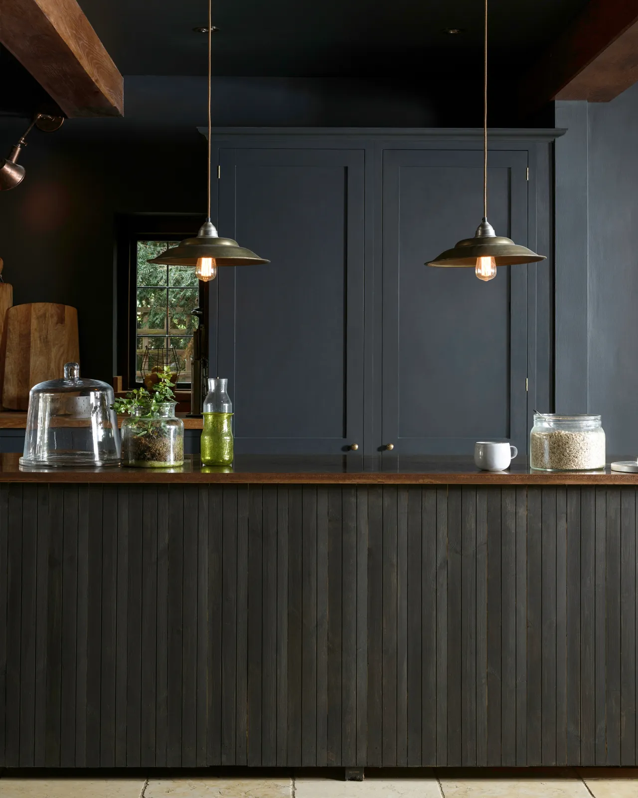 A moody kitchen with dark blue painted ceiling, walls and kitchen pantry cupboard with a vintage slatted island with an aged copper worktop in the middle of the room.