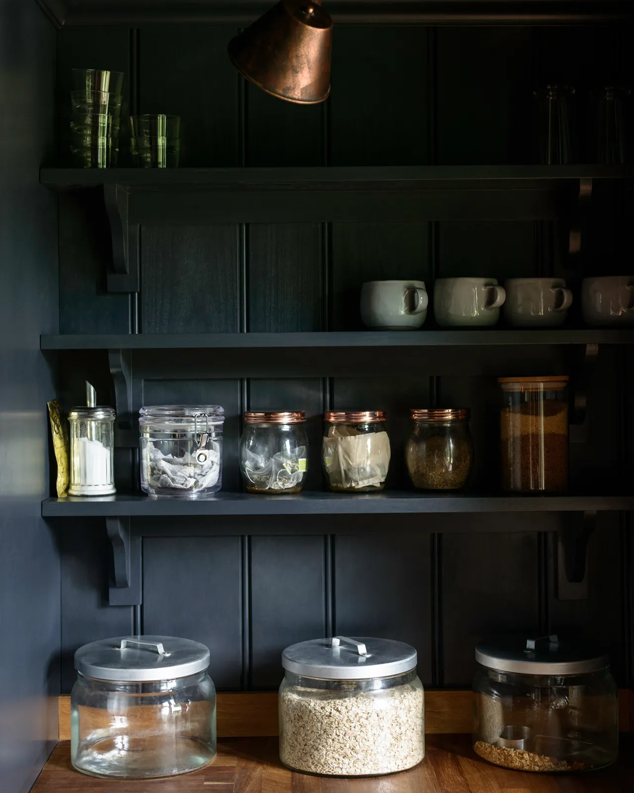 A dark blue painted tongue and groove panelled wall with Shaker-style shelving holding several jars of grains and cereals.