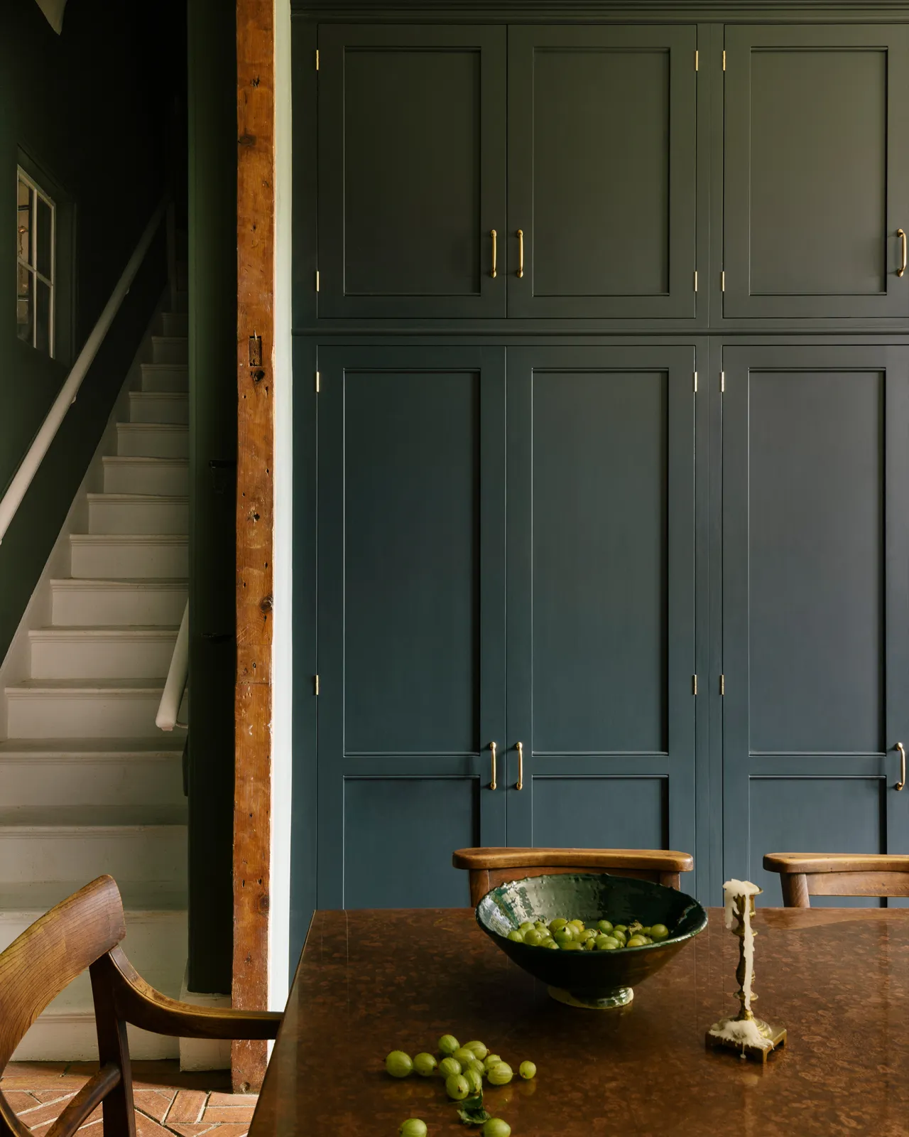 A straight-on view of tall Georgian-style kitchen pantry cupboards painted in a dark blue. There are white stairs to the left of the cupboard and an aged copper tabletop with a bowl of gooseberries in the foreground.