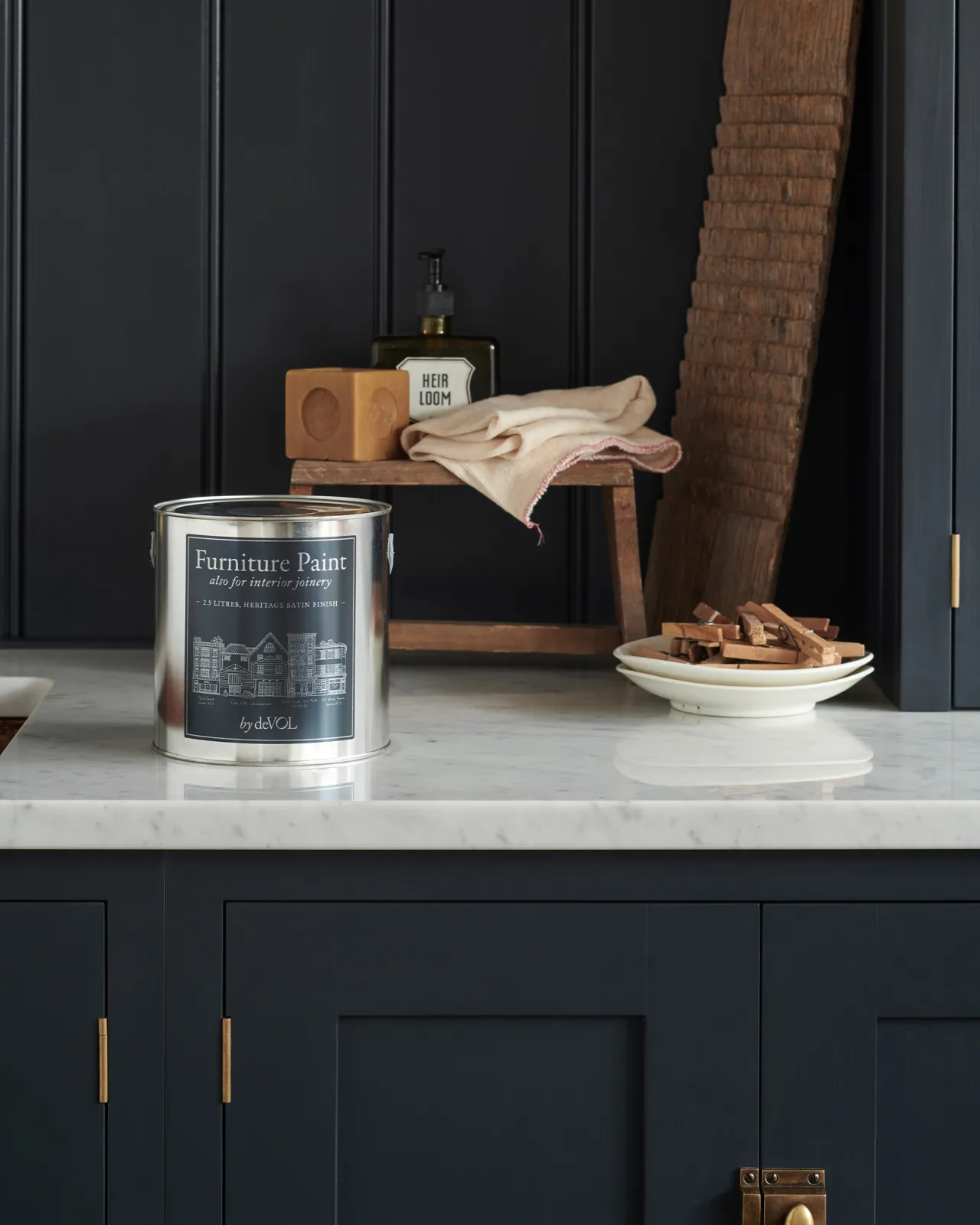 A close-up view of a silver paint tin with a blue label on a Carrara marble worktop, with dark blue painted cupboards and tongue and groove-style panelling behind.