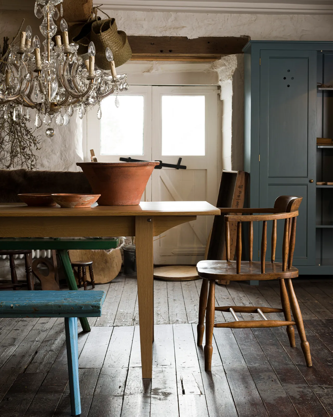 A straight-on view of the end of a mid-century modern-style oak dining table in a rustic cottage with dark worn floorboards and white brick walls.
