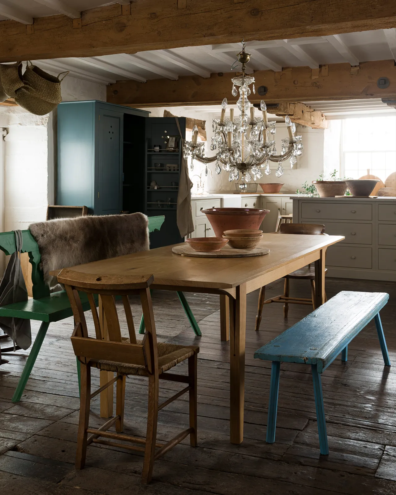 An oblique view of a mid-century modern-style oak dining table in a rustic cottage with dark worn floorboards and white brick walls.