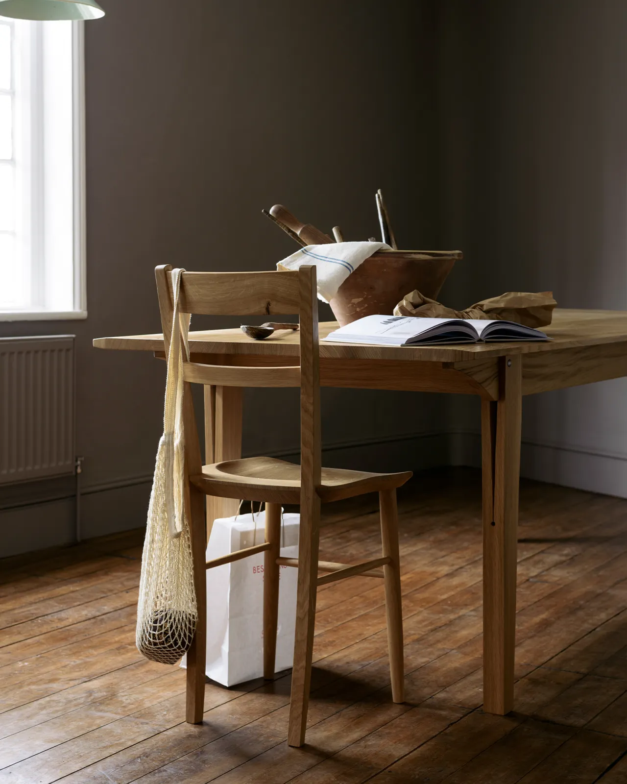 An oblique view of mid-century-style dining chair at the end of a mid-century modern-style oak dining table in a dark room with wooden floorboards.