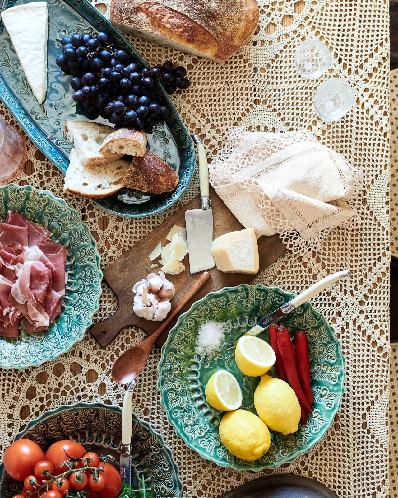 An overhead view of several oval and round handmade bohemian-style ceramic platters, with black grapes, cheeses, homemade bread, lemons, chillis and prosciutto ham on a beautiful crochet lace tablecloth.