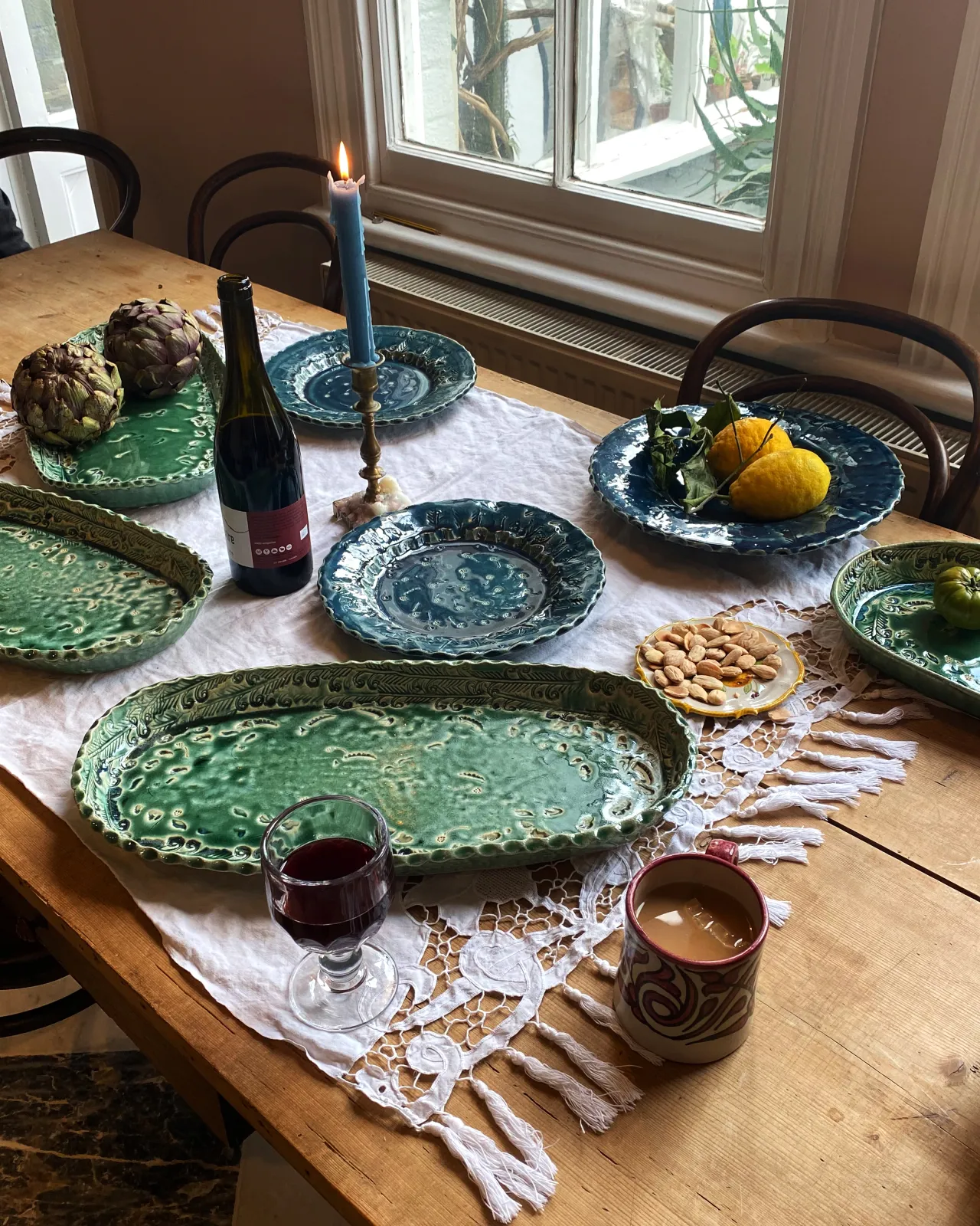 Several oval and round handmade bohemian-style ceramic platters set at a table with a vintage lace tablecloth, a bottle of red wine and a lit candle.