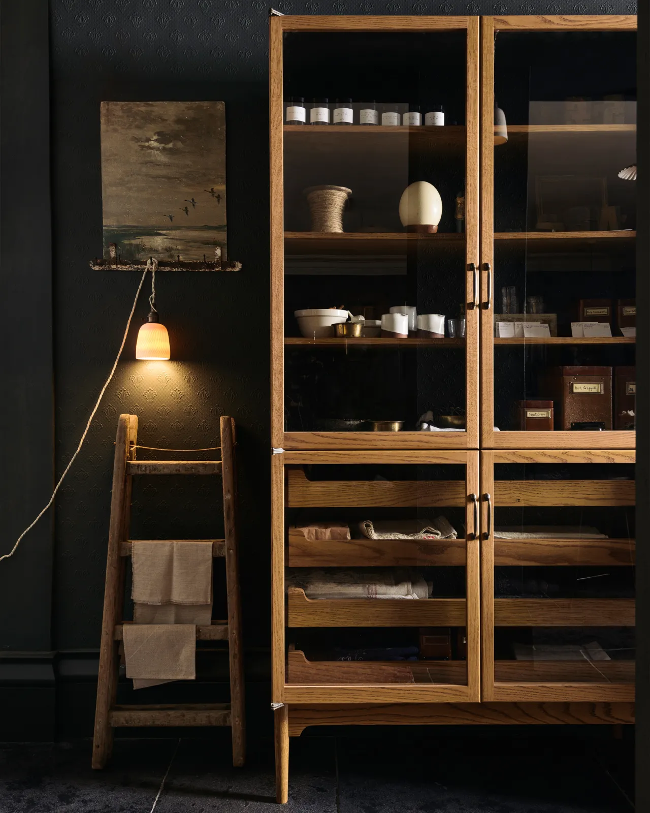 A chattered beaker porcelain plug-in light hanging from wooden ladder beside a mid-century-style glazed pantry cupboard.