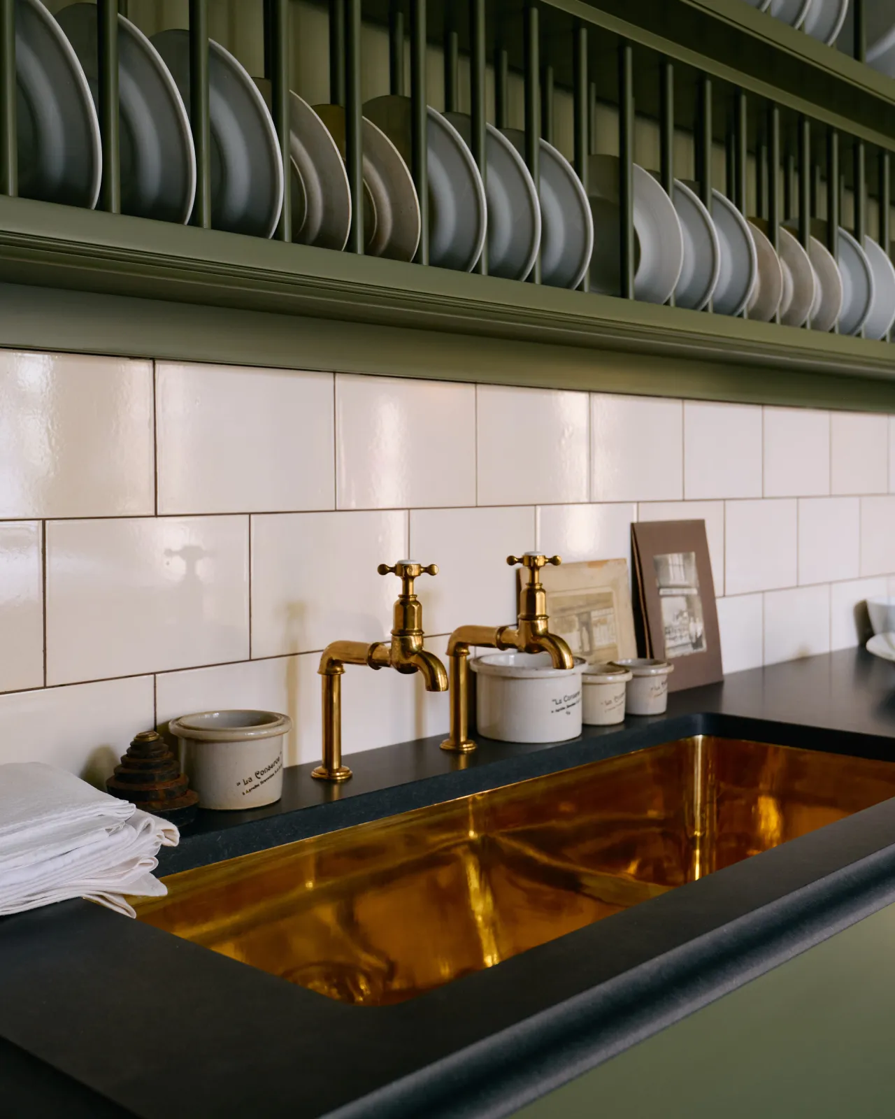 A close-up oblique view of a mossy green painted Victorian-style sink cupboard with a polished brass sink, aged brass bibcock taps and cream subway wall tiles below a traditional wall-mounted plate rack.
