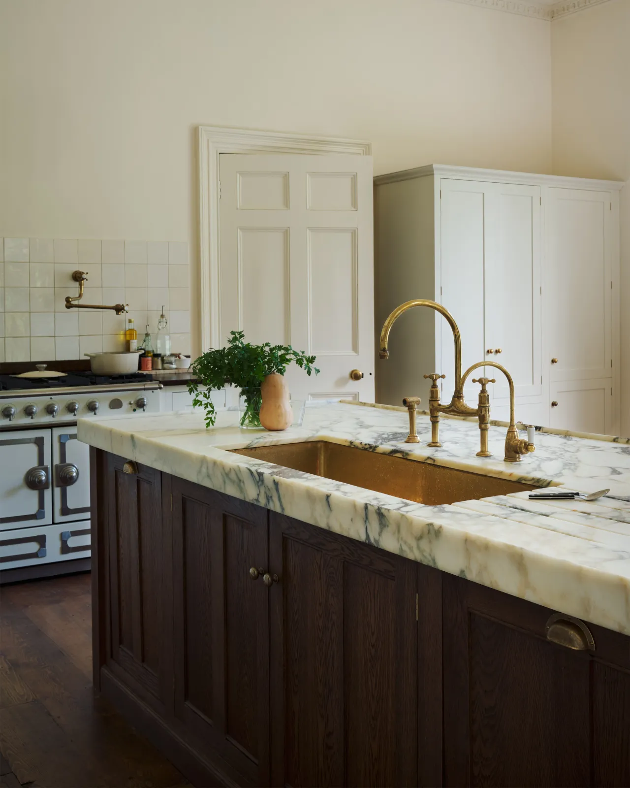 An oblique view of a Georgian-style kitchen with dark stained cabinetry, a deep marble worktop and a large undermounted polished brass sink.