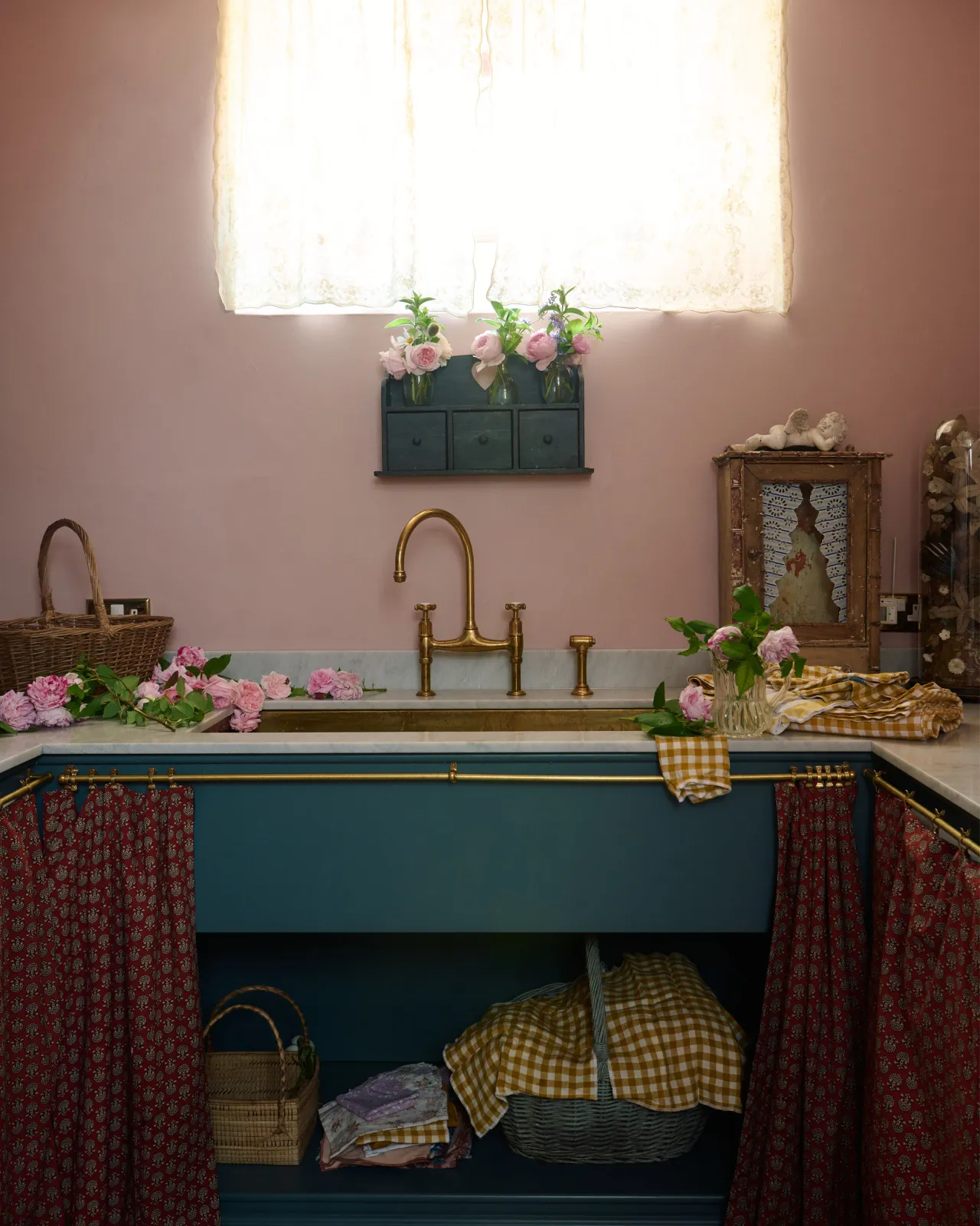 A small and practical Heirloom laundry room features a sink cabinet, a polished brass sink, an aged brass Ionian tap, a curtain rail with crest-patterned red curtains, and shelves. Adorned with eclectic vintage items, pink peonies, tea towels, and wicker baskets, this space creates a romantic and functional atmosphere full of personality.