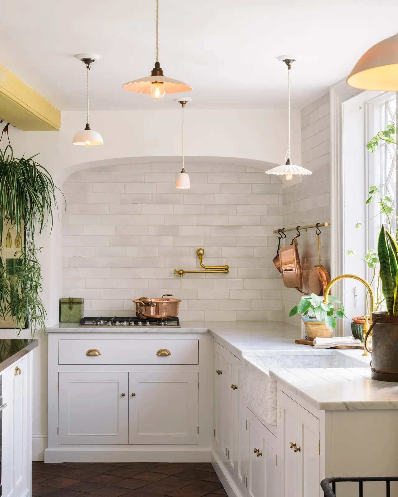 A straight-on view of a Mediterranean-style kitchen with white painted cabinetry, white walls, terracotta flooring and an assortment of unglazed porcelain pendant lights hanging at various heights.