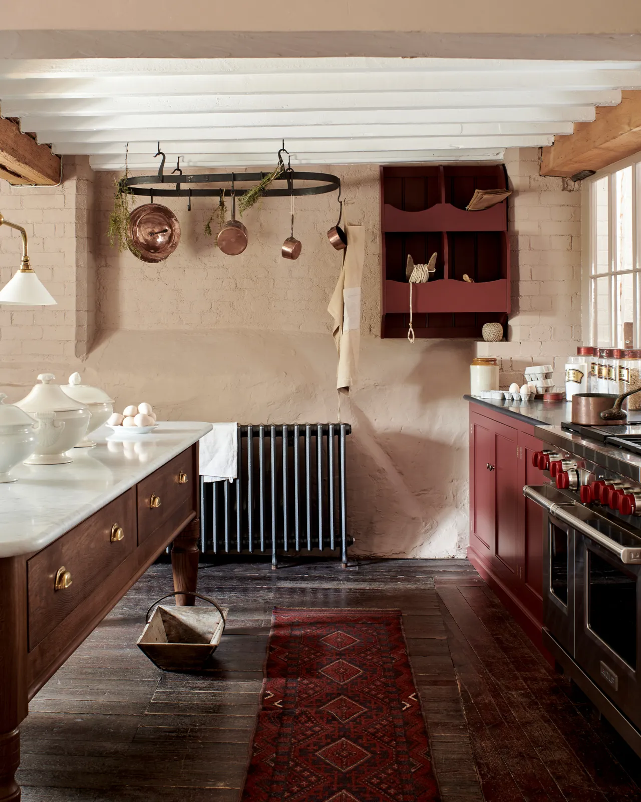 A Victorian-inspired country house kitchen with red painted cabinetry, worn floorboards, pink painted brick walls and a large oval-shaped wrought iron pot hanger suspended from a beamed ceiling via iron hooks.
