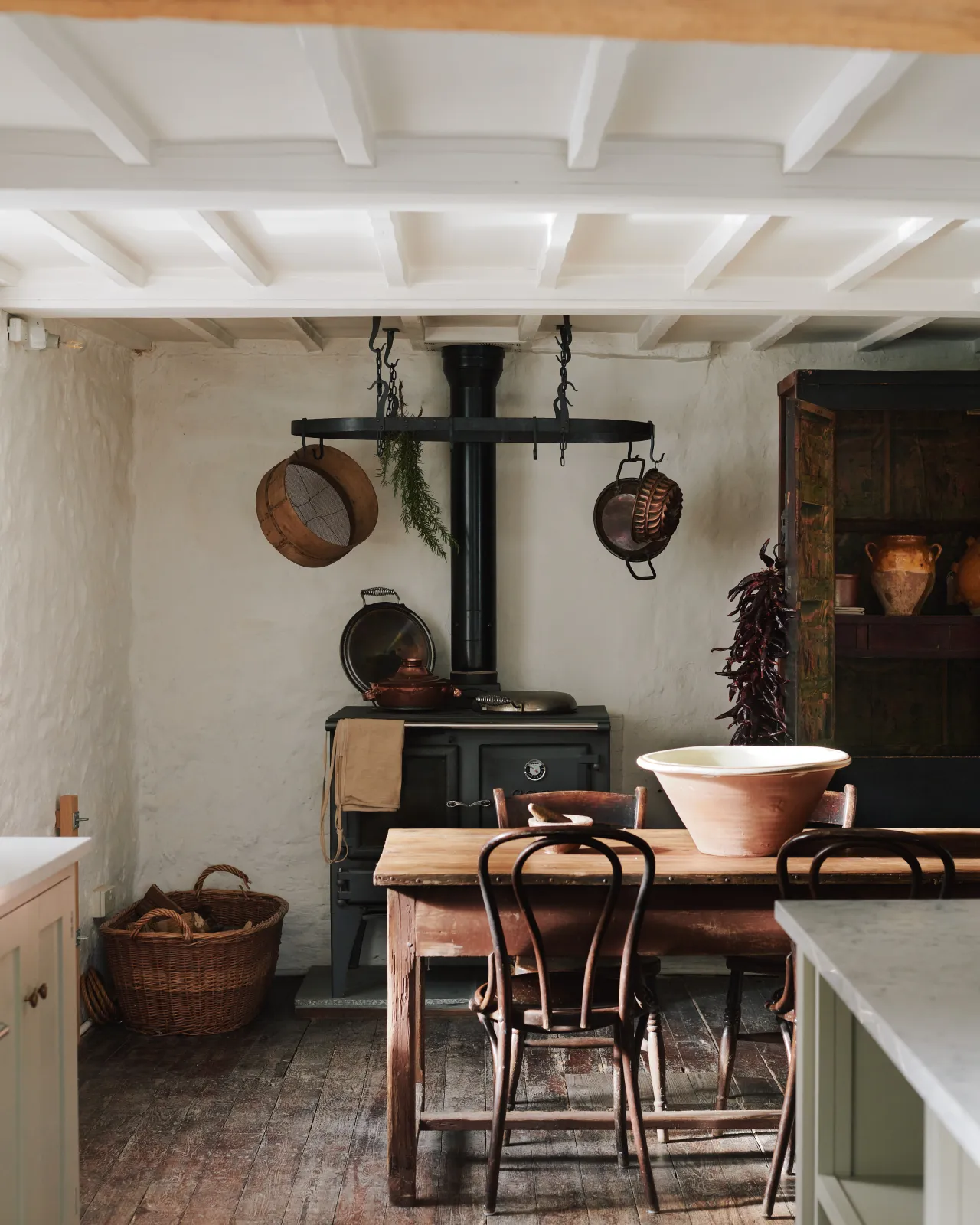 A cottage kitchen dining space with beamed ceilings, old worn floorboards, an antique pine table and chairs and a black Esse stove cooker with a large oval-shaped wrought iron pot hanger suspended from chains above.
