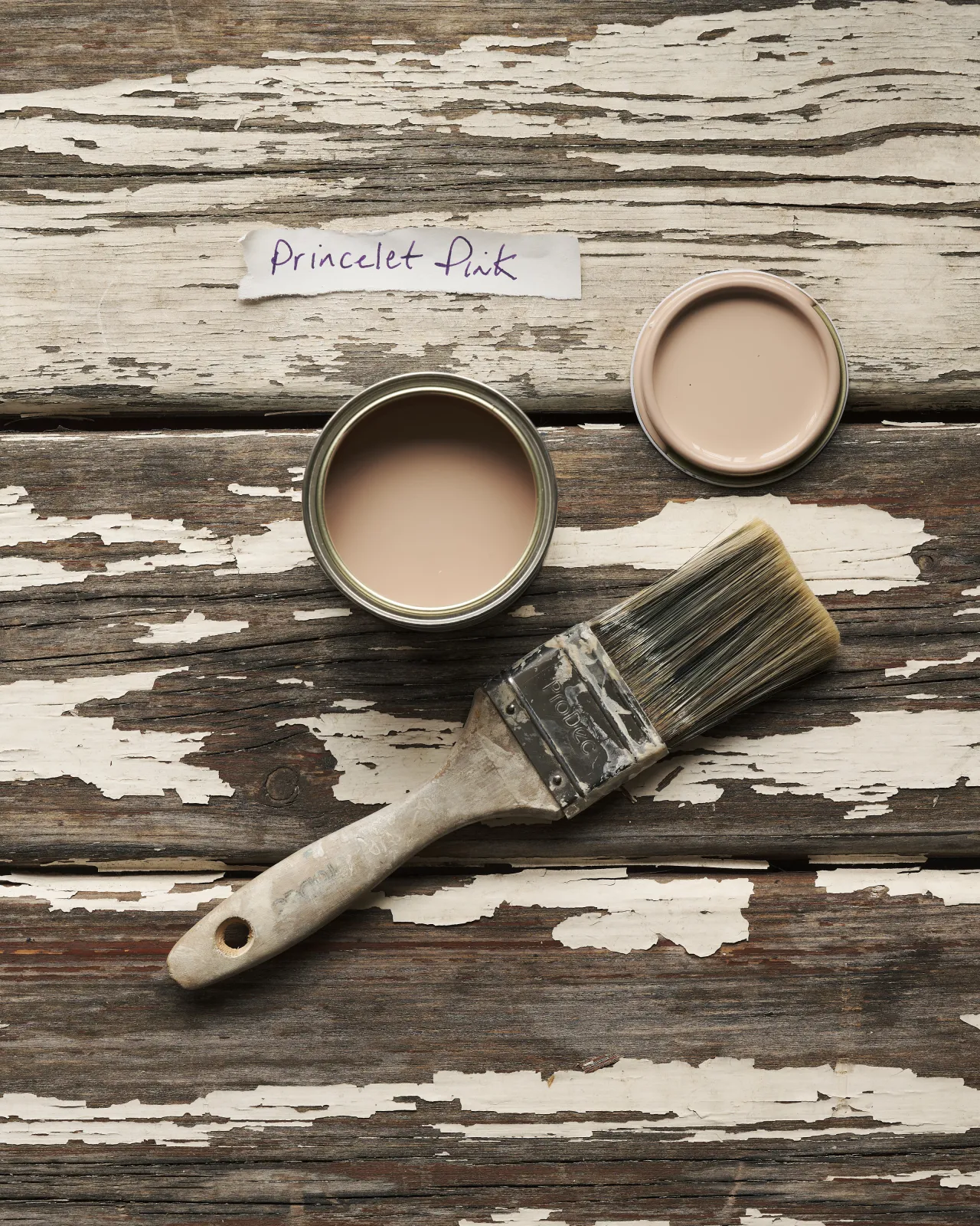 An overhead view of an open paint pot and a worn brush on a rustic tabletop. The soft pink colour inside is labelled 'Princelet Pink'.