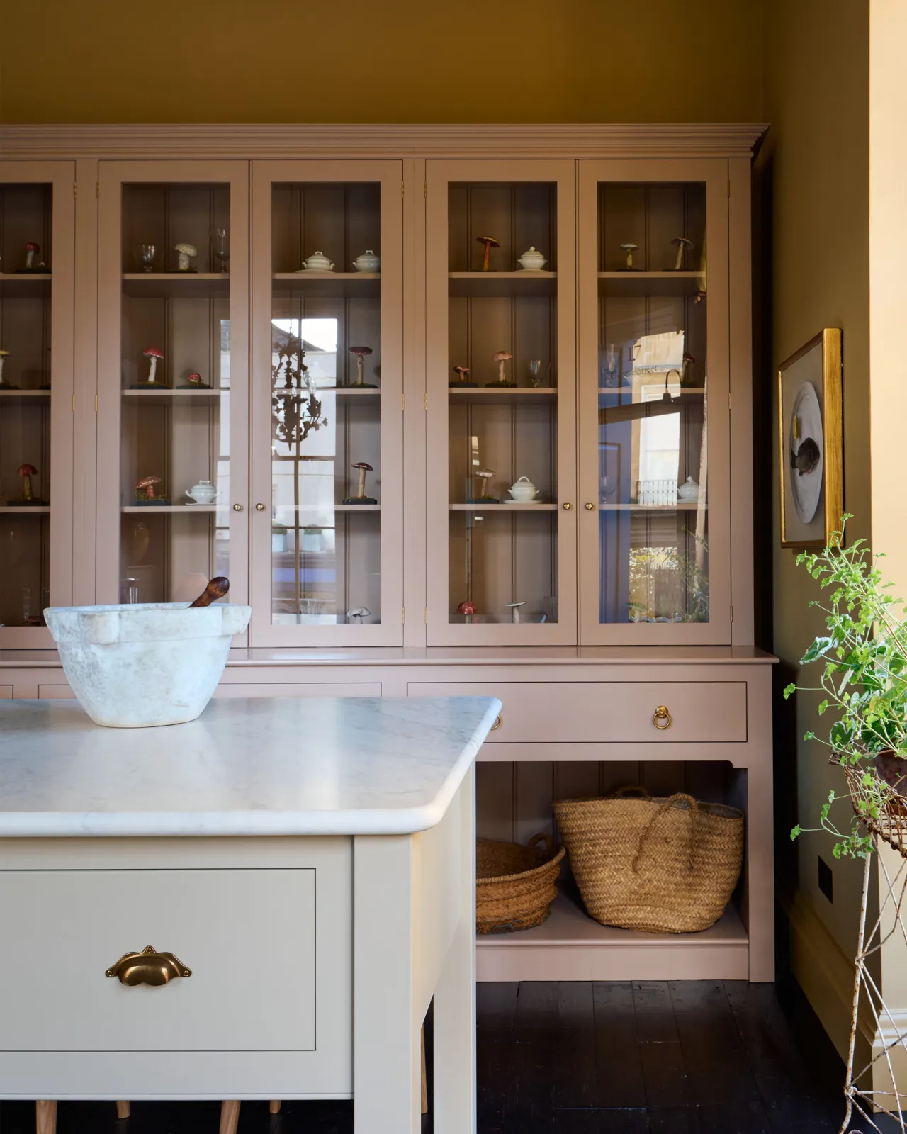 A straight-on view of a tall soft pink painted Georgian-style glazed dresser behind a large soft pink prep table with a Carrara marble worktop.