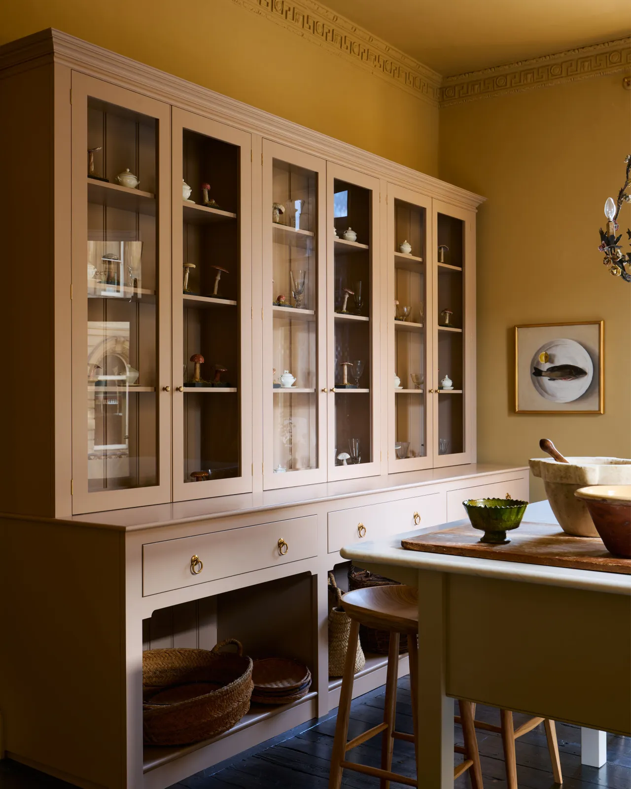 An oblique view of a tall soft pink painted Georgian-style glazed dresser in an atmospheric room with warm yellow walls.