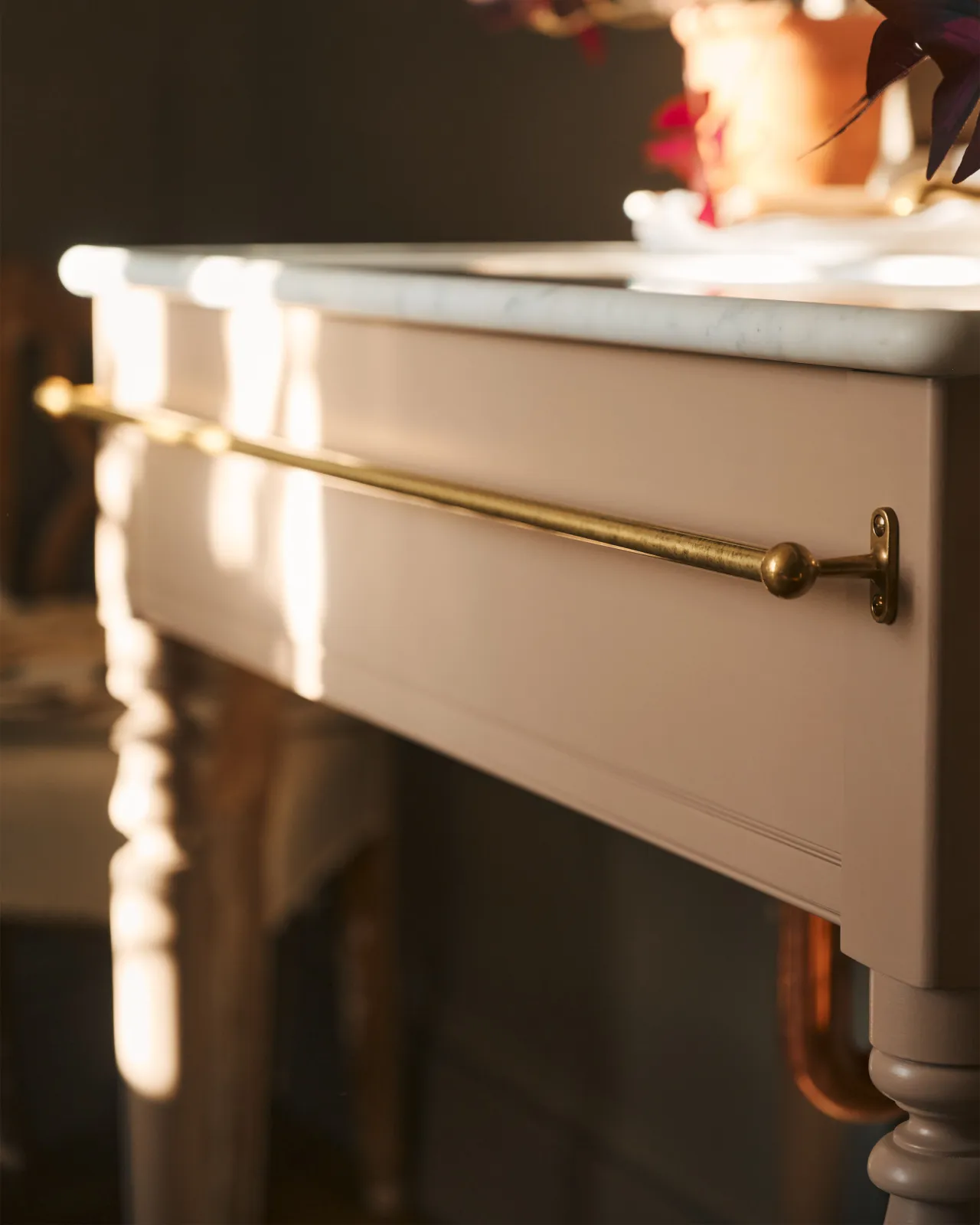 A detailed view of an aged brass towel rail attached to the front of a soft pink painted Victorian-inspired washstand.