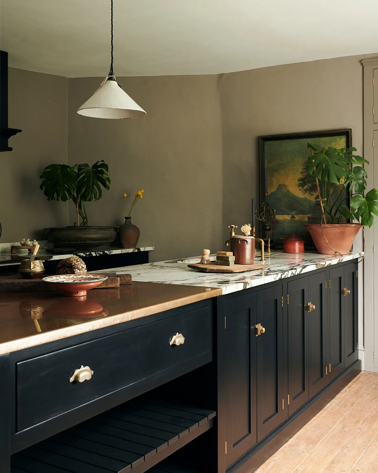 An oblique view of a black painted Shaker-style kitchen peninsula with brass hardware, aged copper and dramatic quartz worktops, grey walls and a large vintage oil painting.