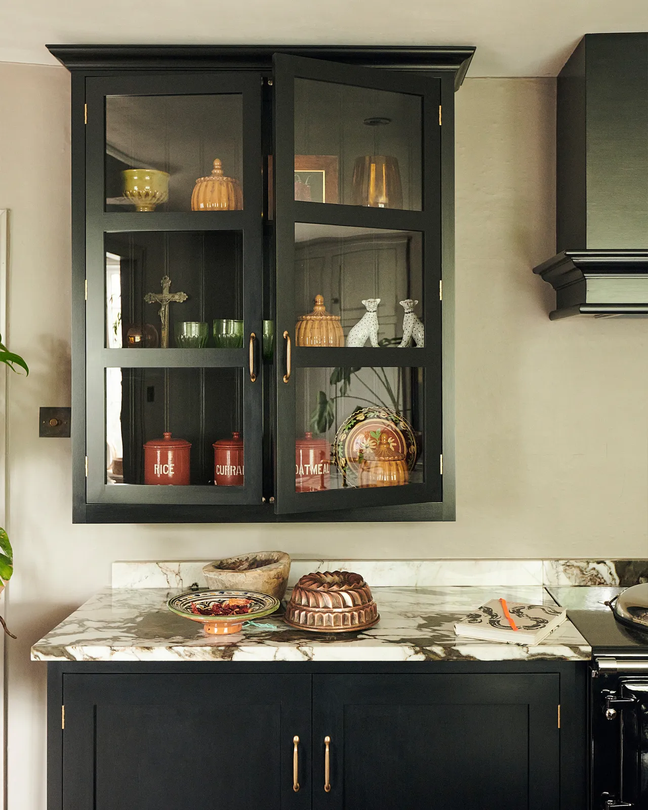 A straight-on view of a Shaker-style glazed wall cupboard painted black inside and out with several red-brown jars and pots inside.