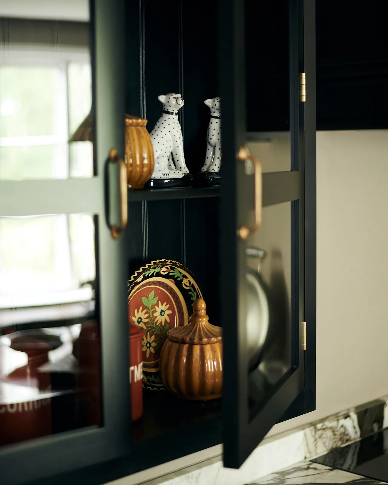A close-up oblique view of a Shaker-style glazed wall cupboard painted black inside and out with one door open and several painted porcelain dog statues inside.