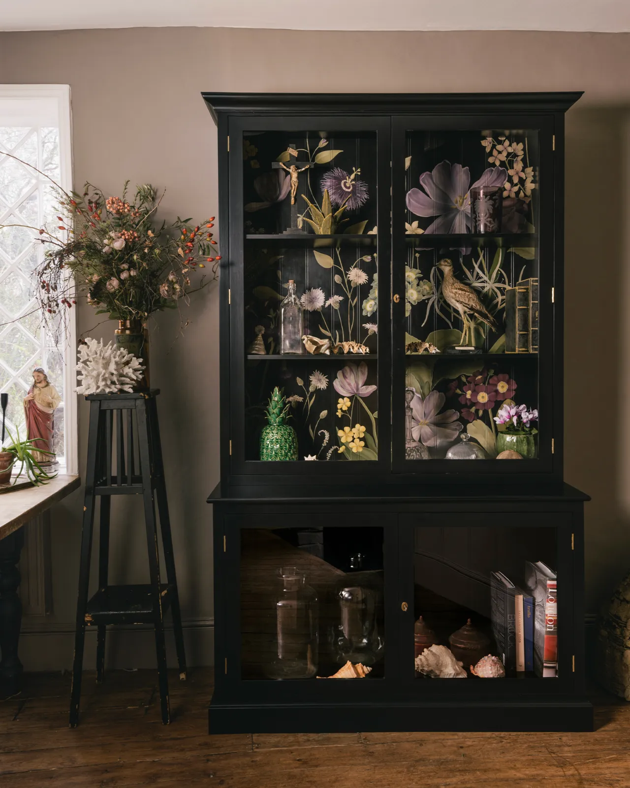 A tall museum-style display cabinet with glazed doors, painted black inside and out and with an elegant and bustling painted floral design on the tongue and groove back panel.