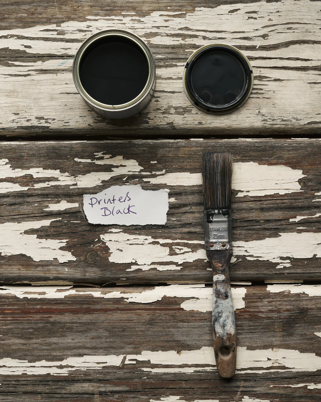 An overhead view of an open paint pot and a worn brush on a rustic tabletop. The black colour inside is labelled 'Printer's Black'.