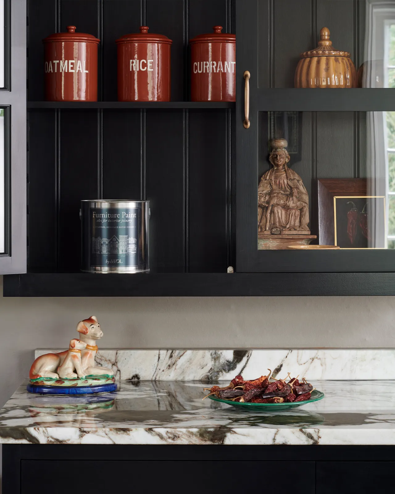 A close-up straight-on view of a Shaker-style glazed wall cupboard painted black inside and out with one door open and several red-brown jars and pots inside.