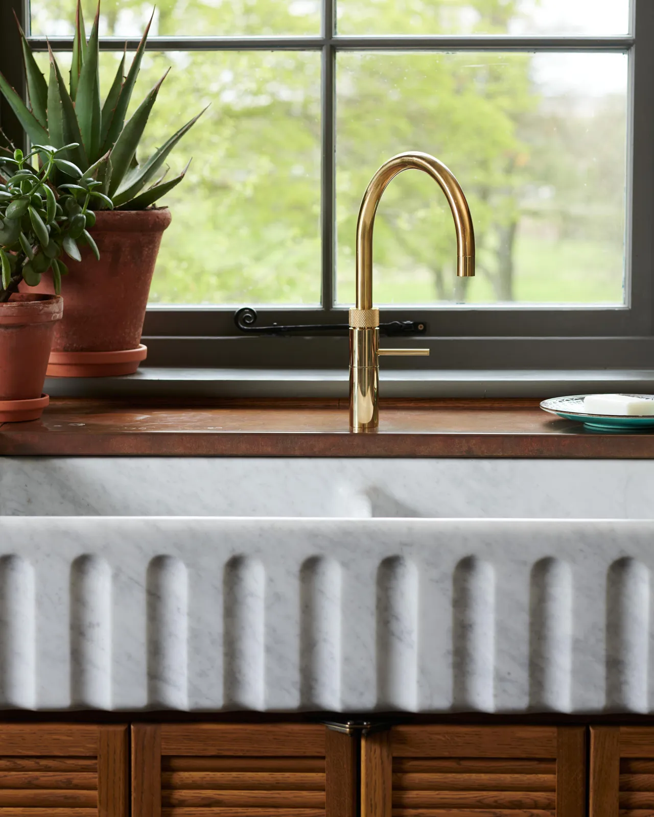 A close-up view of a contemporary-style brass tap above a grand fluted double bowl Carrara marble sink and an aged copper worktop.