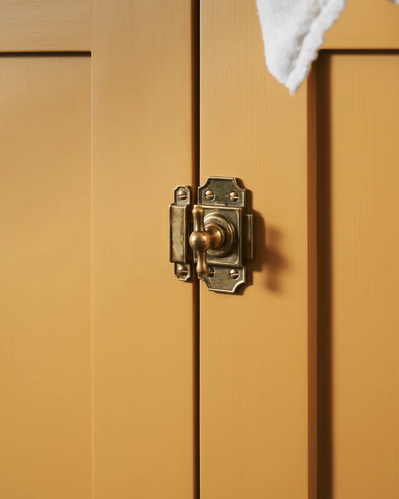 A close-up oblique view of a Victorian-inspired rack and pinion cupboard catch in a warm burnished brass finish, fitted to a yellow painted door.