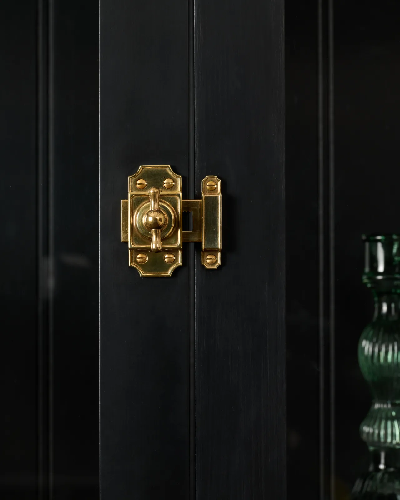 A close-up straight-on view of a Victorian-inspired rack and pinion cupboard catch in a warm burnished brass finish, fitted to a black painted door.