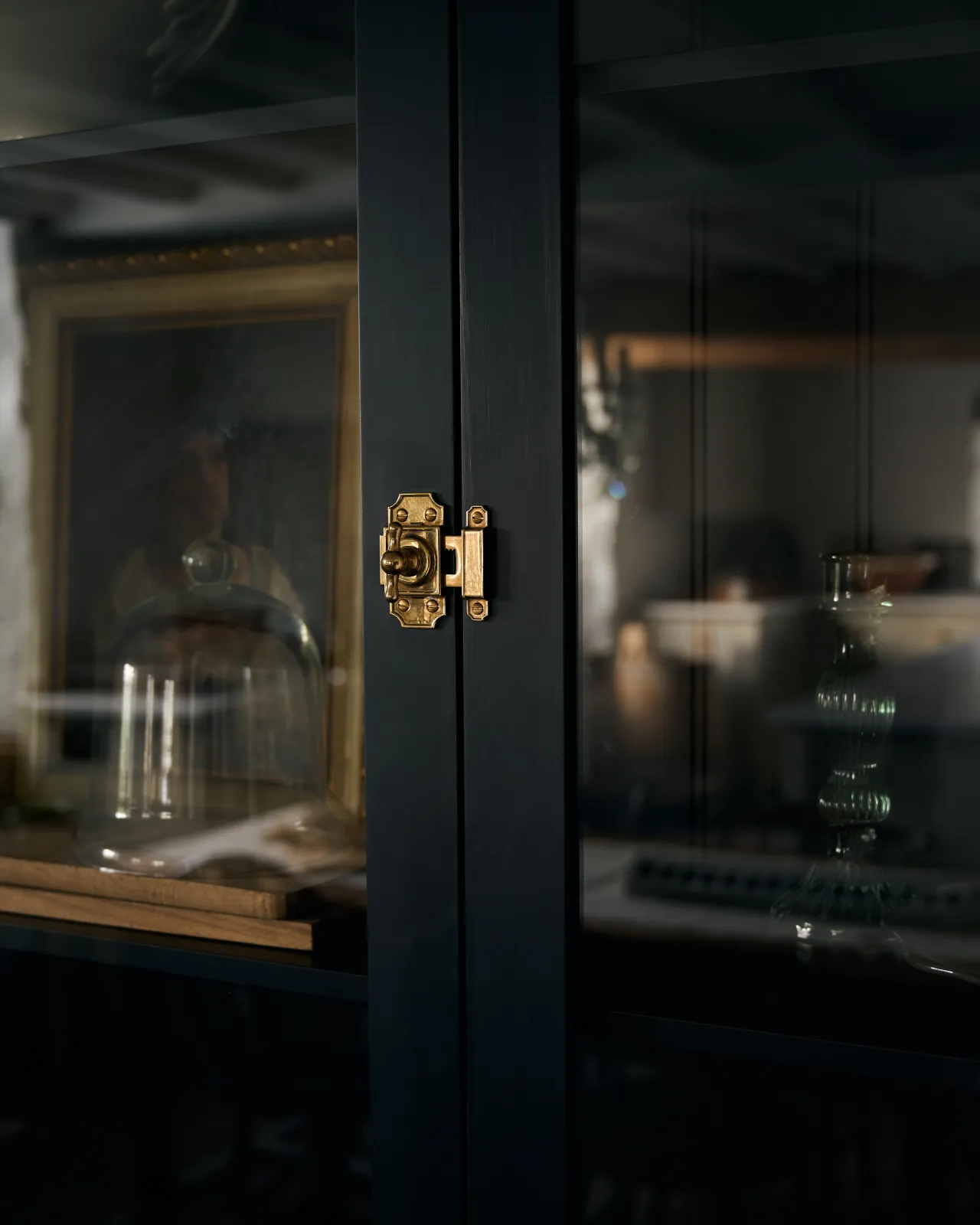 An oblique view of a Victorian-inspired rack and pinion cupboard catch in a warm burnished brass finish, fitted to a tall glazed cupboard painted black inside and out.
