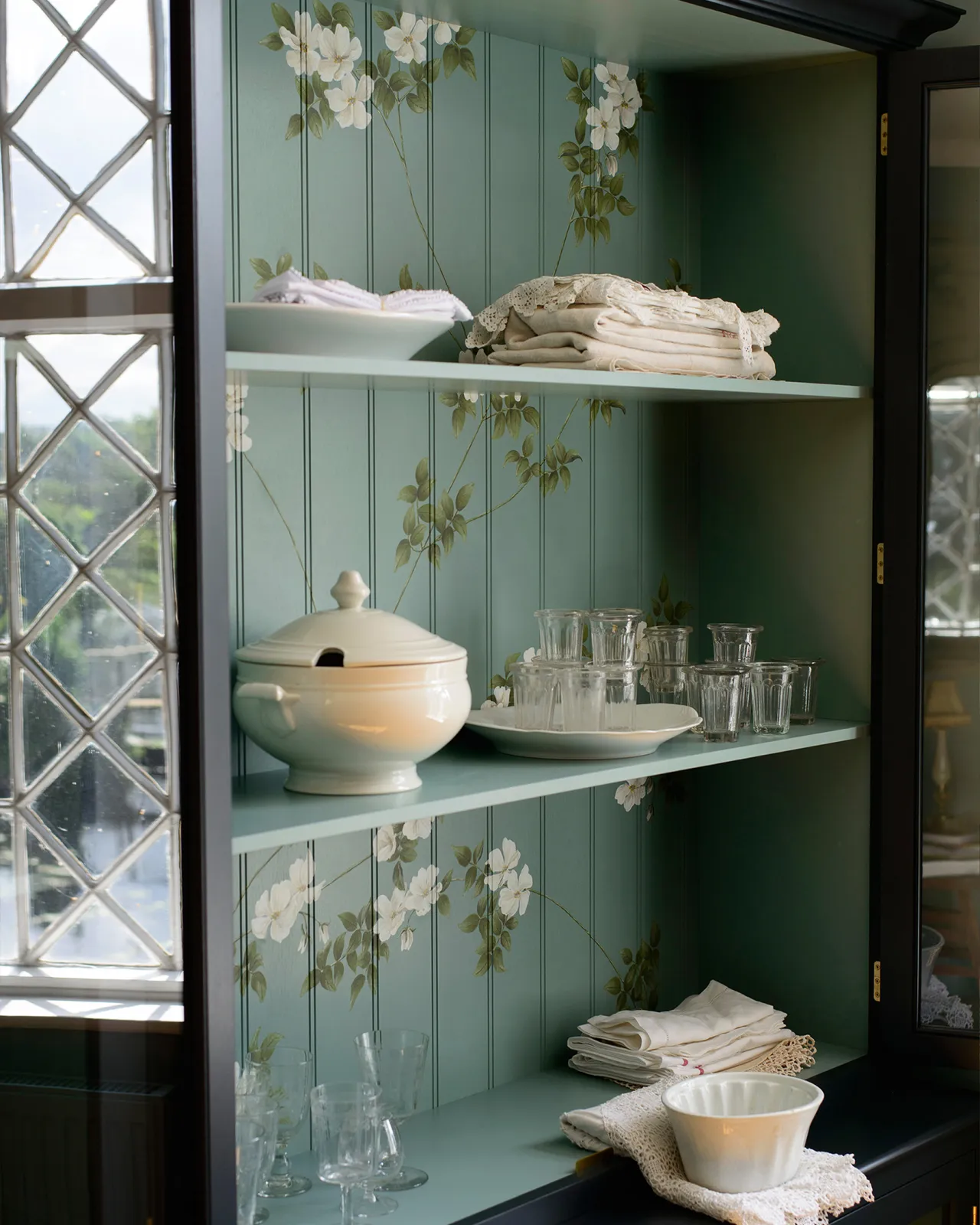 An oblique view of the interior of a display cabinet with a duck-egg blue interior and painted flowing white dog-roses on the tongue and groove back panel.