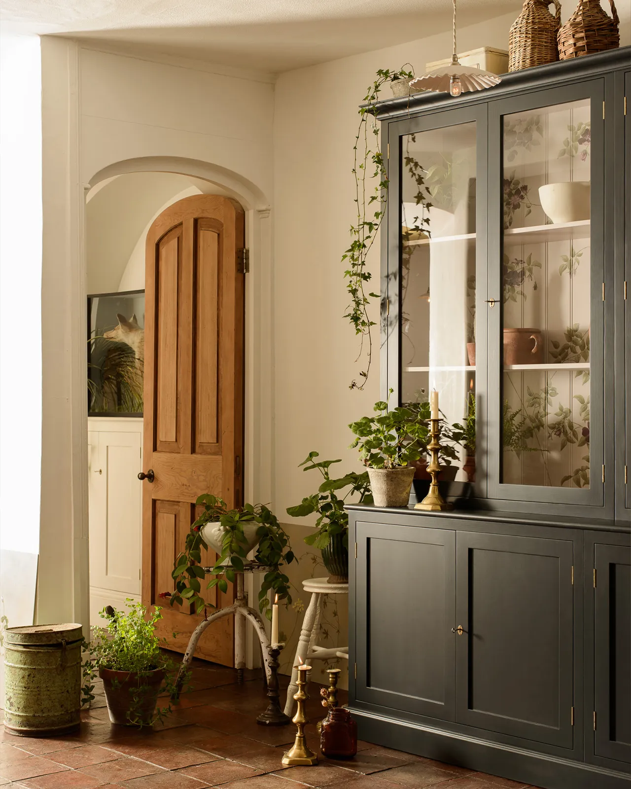 An oblique view of a tall double museum-style display cabinet with glazed doors, painted a dark blue outside with a light pink interior and painted flowing purple dog-roses on the tongue and groove back panel.