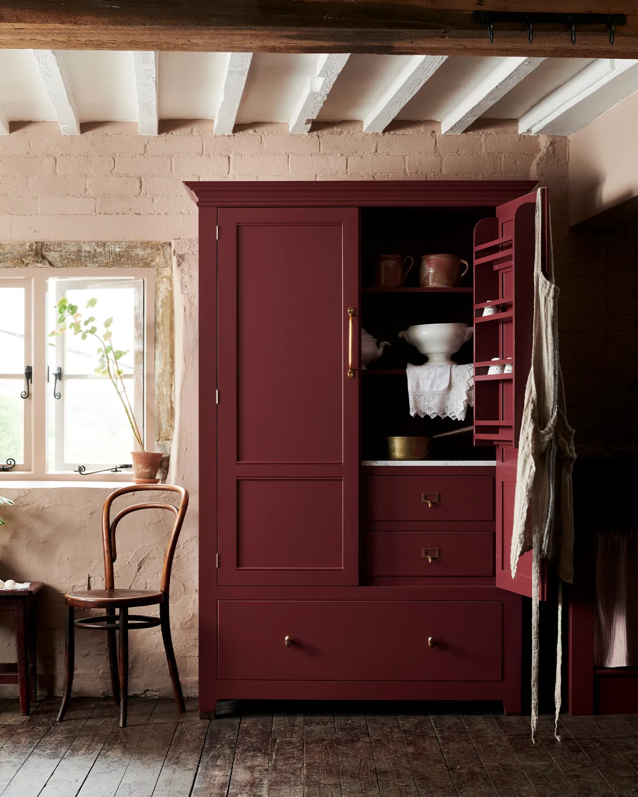 A red painted Georgian-style pantry cupboard with brass hardware and one door open, revealing white vintage crockery inside.