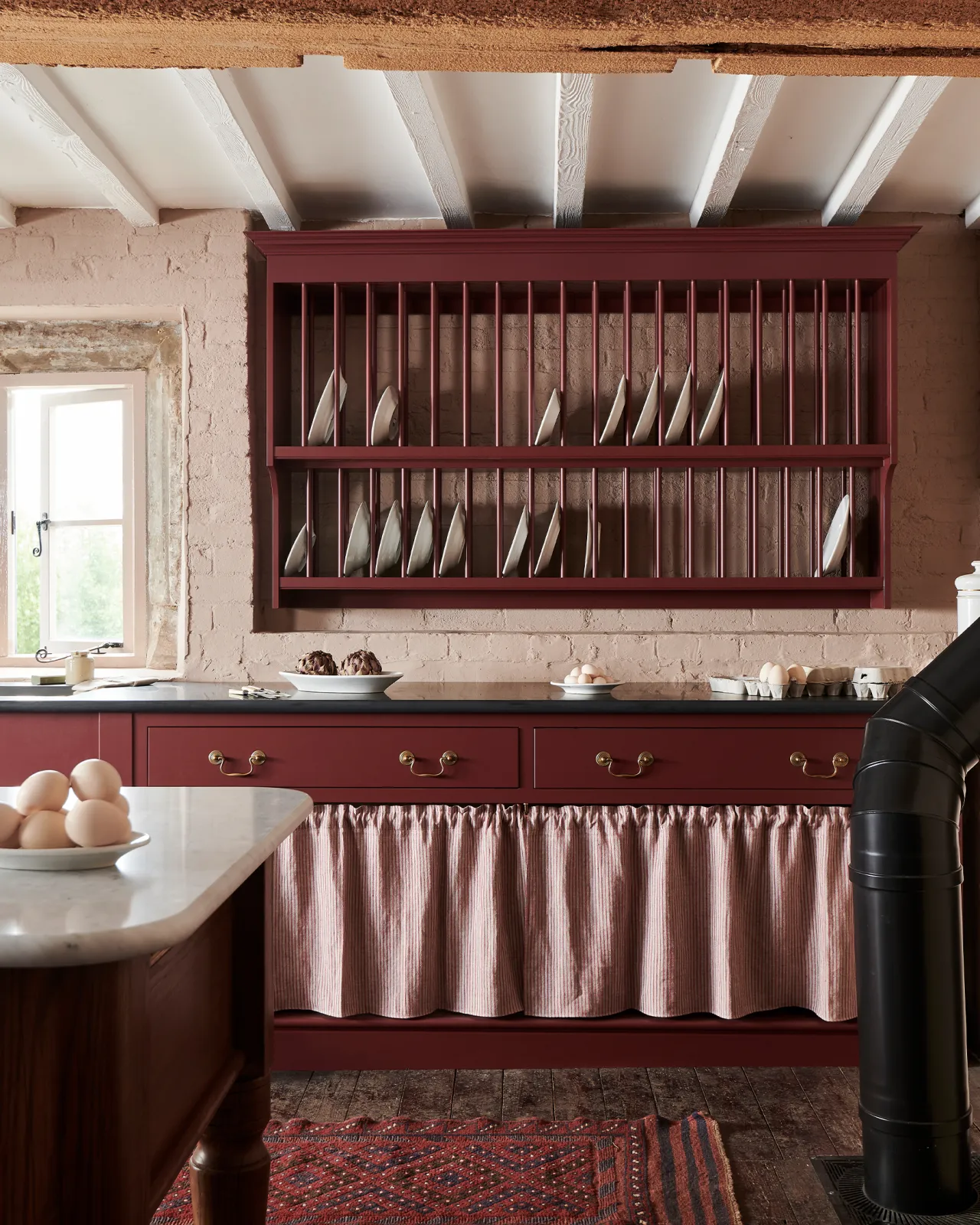 A straight-on view of a red painted Victorian-style kitchen with a traditional wall-mounted plate rack and wide drawers with open storage below.