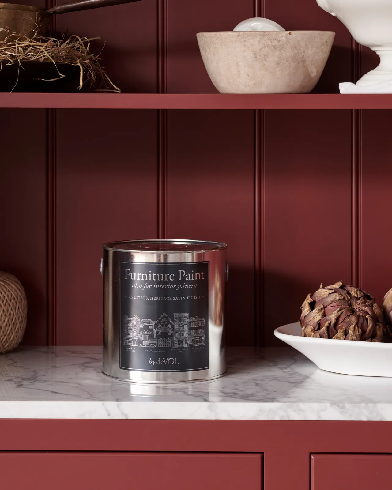 A close-up view of a silver paint tin with a blue label on a Carrara marble worktop, with rich red painted cupboards and tongue and groove-style panelling behind.