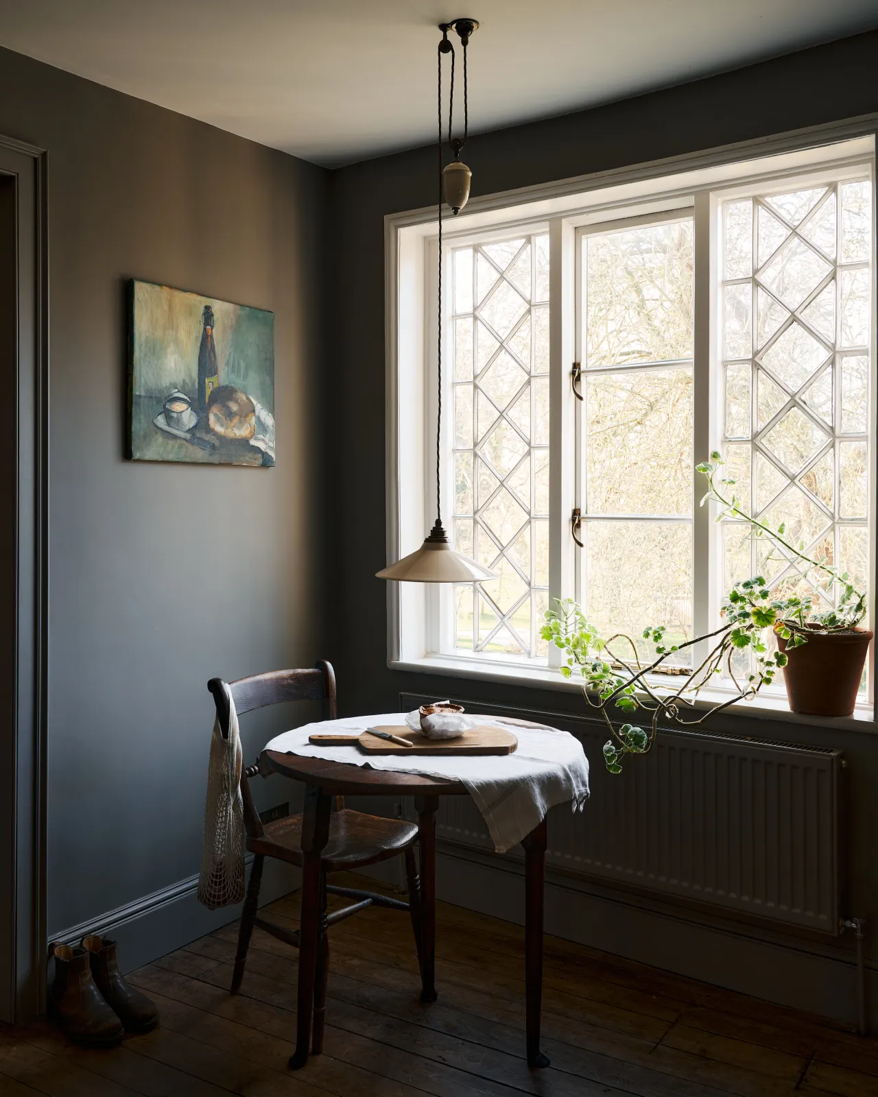 An oblique view of a rise and fall light with a small cream glazed ceramic shade, aged brass fittings and brown twisted fabric flex cord hanging in a dark room by a large Victorian leaded window.