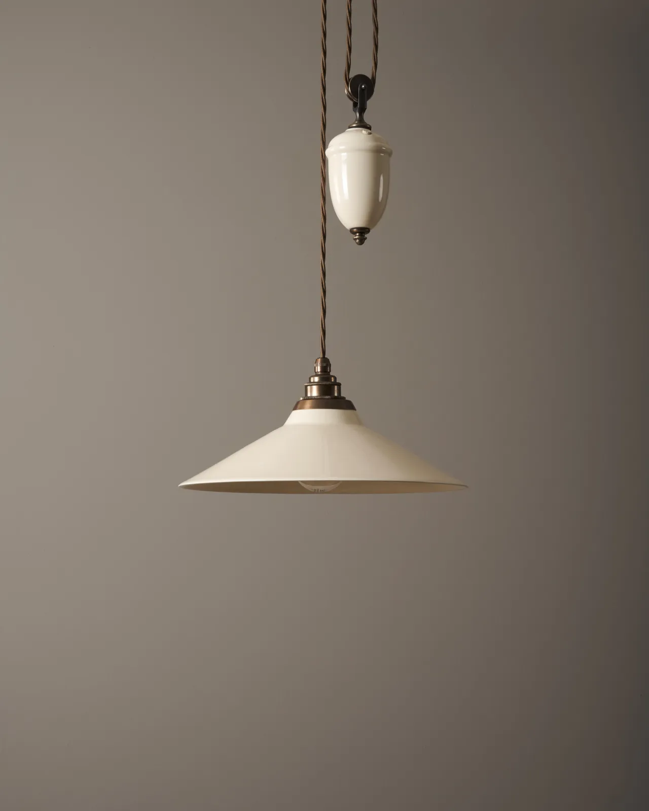 A rise and fall light with a small cream glazed ceramic shade and counterweight, an aged brass E27 lampholder and brown twisted fabric flex cord, the light is turned off.