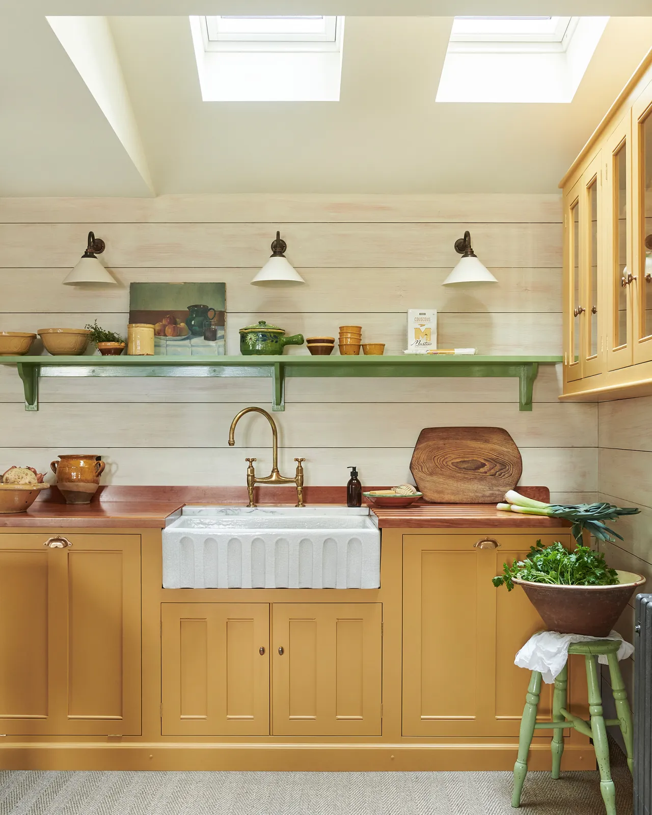 A straight-on view of a yellow painted Georgian-style kitchen sink cabinet in a small cottage kitchen with white painted panelled walls, green shelving and grey carpet flooring.