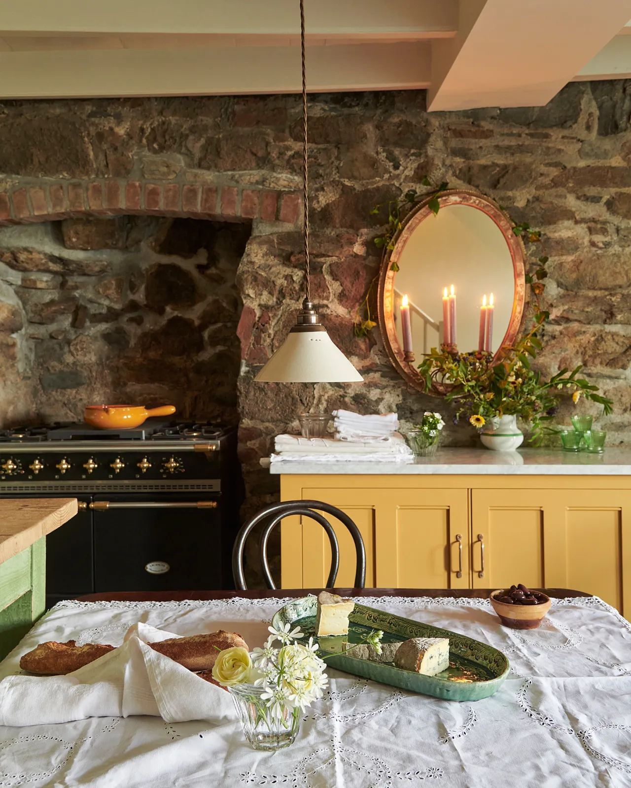 Looking over a table with a vintage lace tablecloth and bohemian ceramic platter at yellow painted Georgian-style cupboards either side of a black range cooker. A stone wall with vintage mirror are seen above.