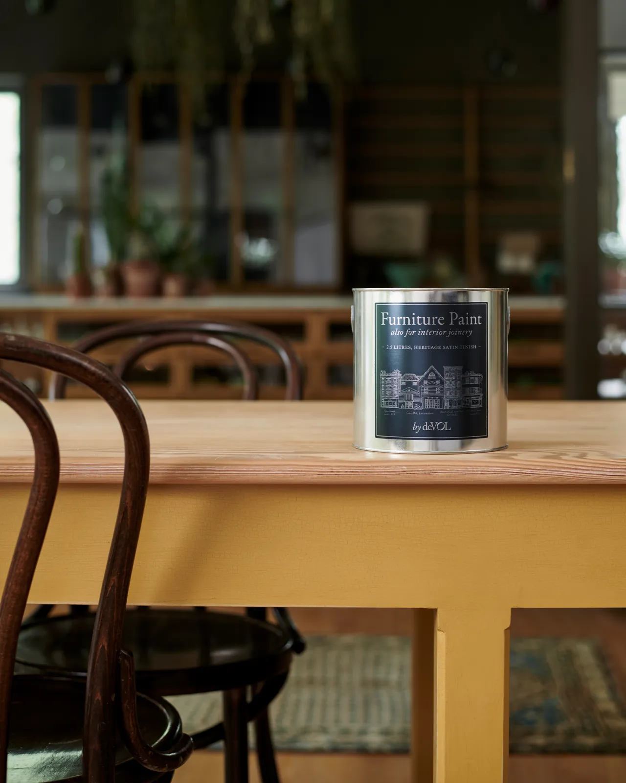 A close-up view of a silver paint tin with a blue label on a yellow painted prep table with a smooth pine tabletop.
